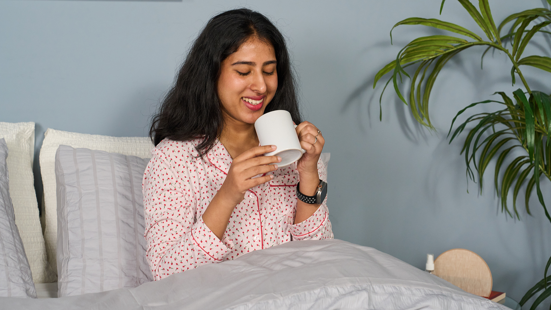 A woman sits up in bed and drinks from a mug