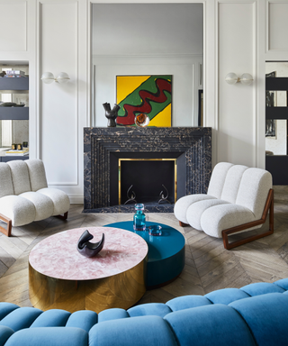 Modern lounge space with curved tufted blue couch, white tufted armchair, round terrazzo coffee table central to fireplace with mirror above it reflecting bright artwork