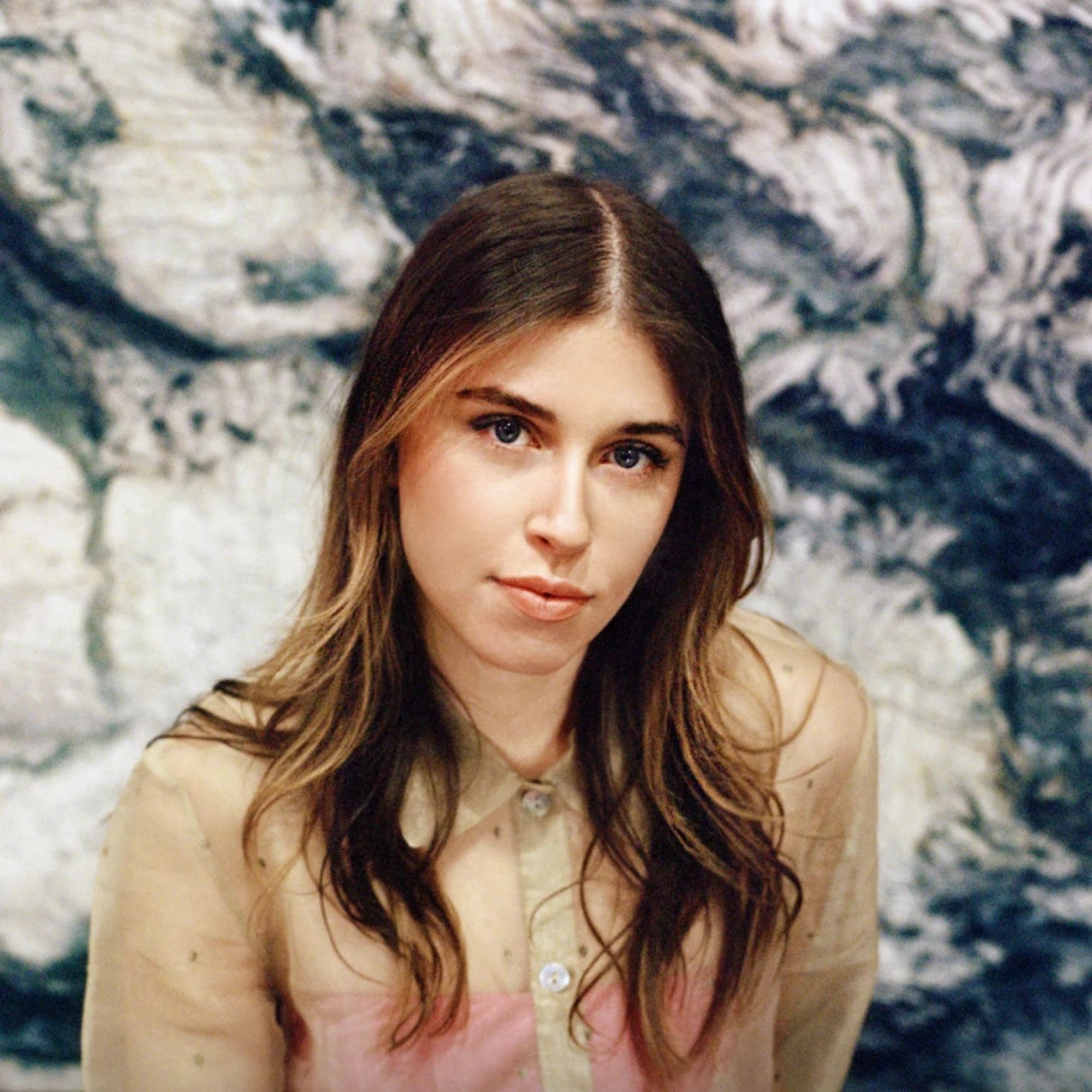 woman with long brown hair and sheer shirt in front of stone slab looking at camera