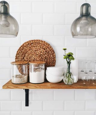 A white tiled kitchen wall with floating wooden shelf. Decanted dried food goods on the shelf with a place mat
