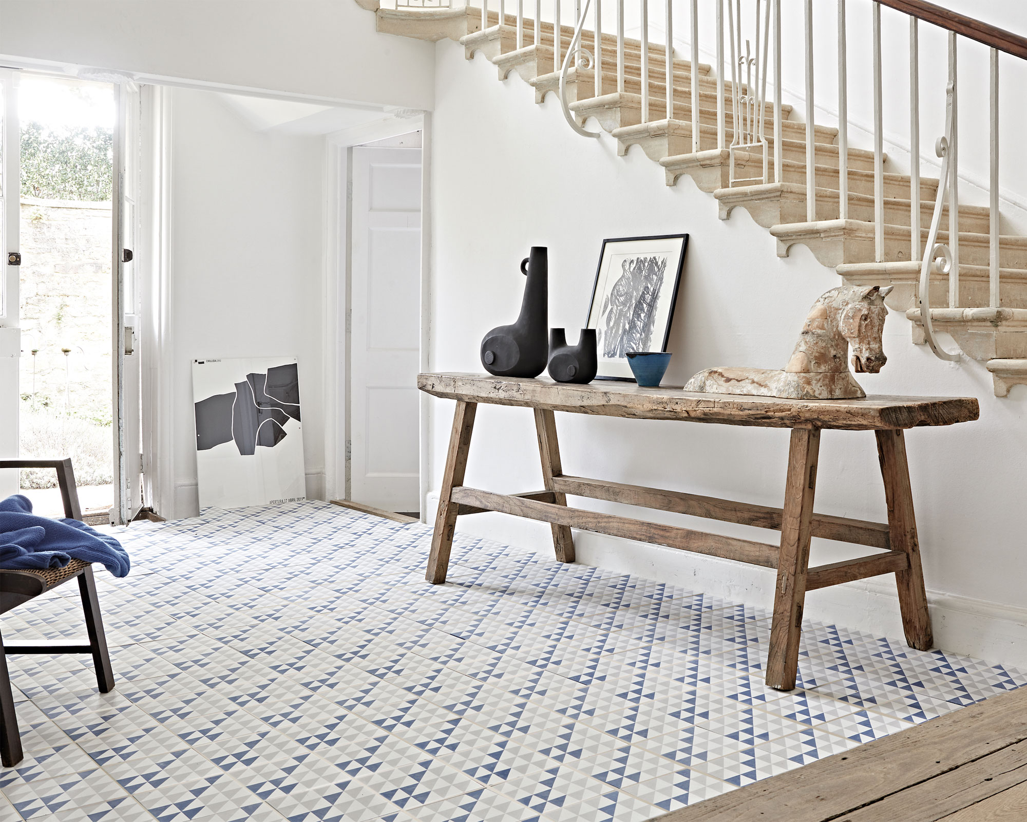 Country-cottage style hallway with large stone staircase, wooden flooring, lower level flooring in gray and blue geometric, triangle tiles, unique dark wooden console table decorated in objects, white painted walls