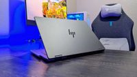 A sleek laptop with a metallic finish is on a wooden desk, partially open. A gaming chair and monitor displaying a colorful landscape are in the background.