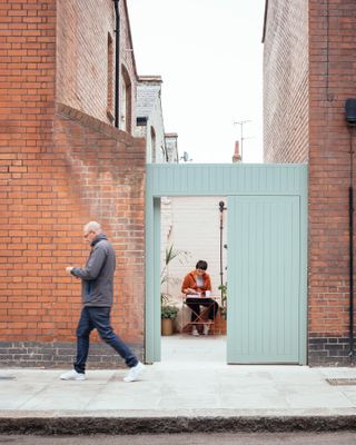 Man walk on pavement past open door to court yard where a lady is sitting and filling out a form.