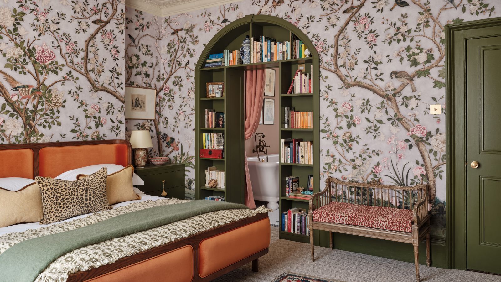 Cozy bedroom with tree and leaf wallpaper, dark khaki green painted door and woodwork, including a built-in arched book shelf storage leading into a bathroom with freestanding tub visible. The bed has a mid century orange velvet header and footer and there are textured pillows and throws on the bed.