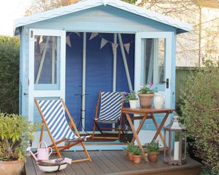 Coastal inspired shed with soft blue exterior, and bolder blue interior, styled with pretty bunting and diagonal stripe deckchairs.