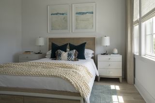 white bedroom with linen drapes and blinds, wooden floor, artwork, cream knitted blanket, wooden bed, white bedside tables, rug, matching table lamps