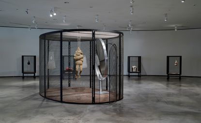 An exhibition space featuring a circular metal cage with a character hanging from its roof and a large circular mirror. 