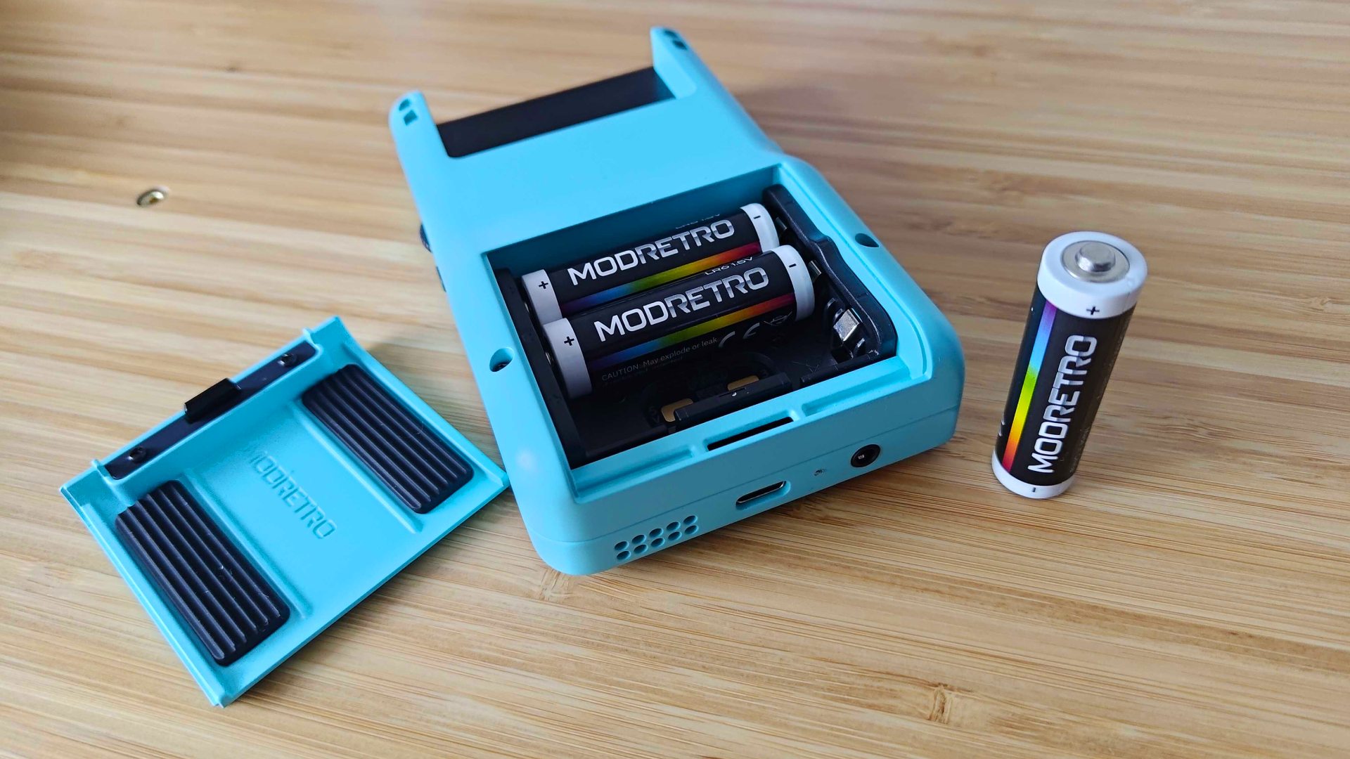 Modretro Chromatic lying face down on woodgrain surface with battery compartment open and one AA cell removed.