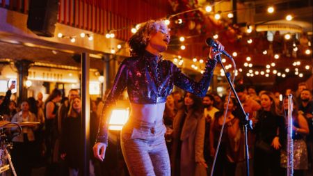 an image of a woman singing into a mic on stage at an event which has lots of festoon lighting around and a crowd looking on
