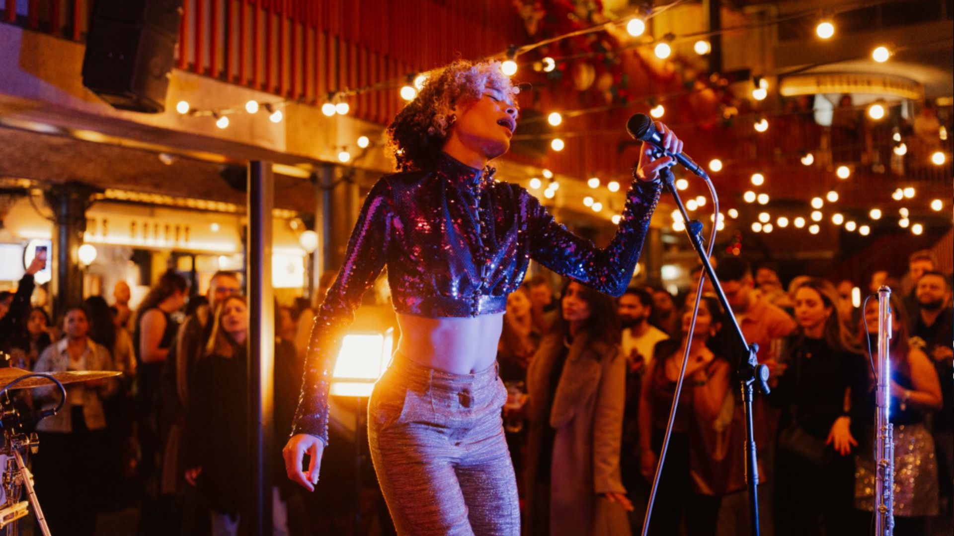 an image of a woman singing into a mic on stage at an event which has lots of festoon lighting around and a crowd looking on