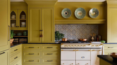 a kitchen with olive oil colored cabinetry, a butter yellow stove, black countertops, and a decorative backsplash. Above the stove hangs three decorative plates