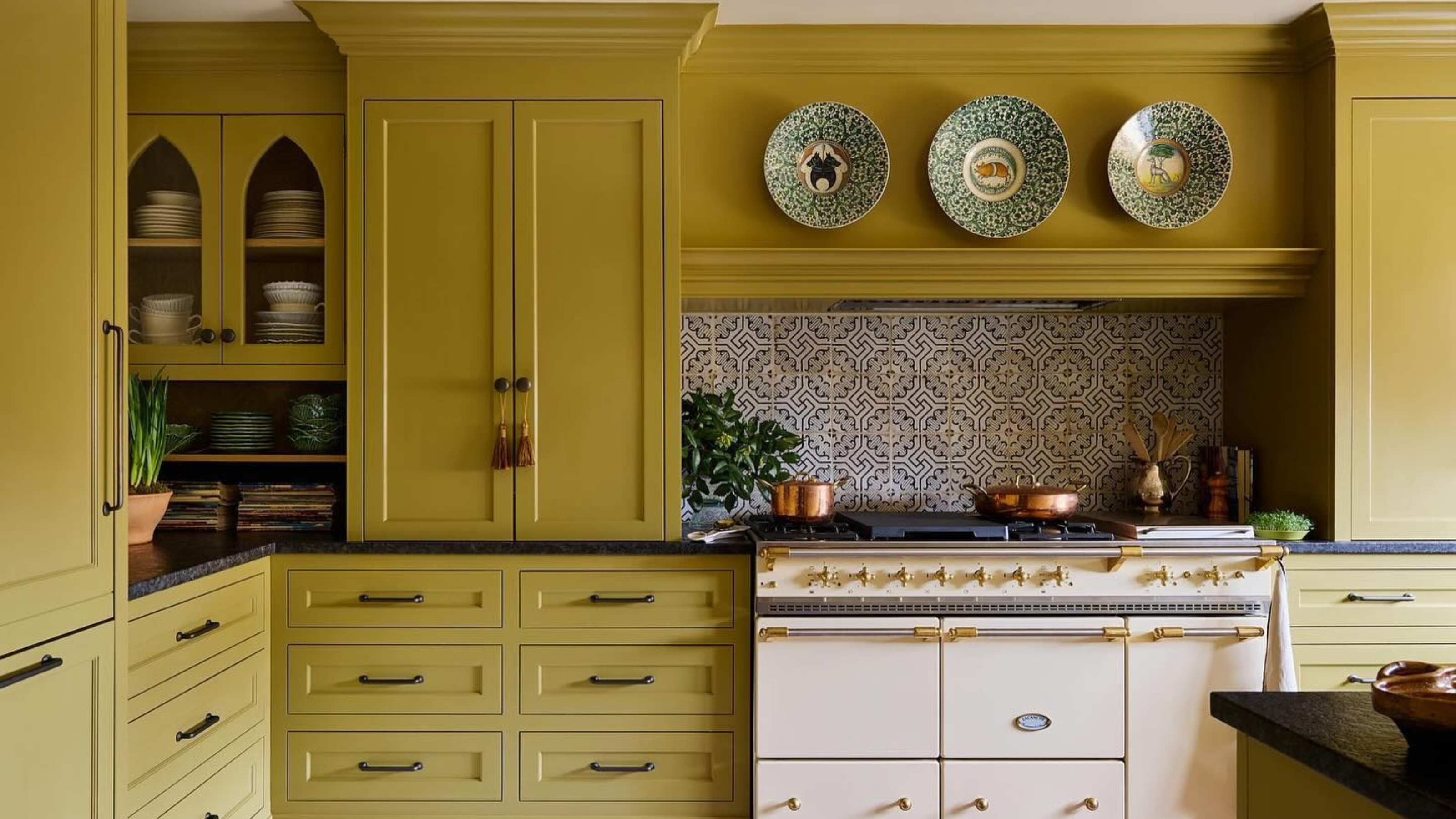 a kitchen with olive oil colored cabinetry, a butter yellow stove, black countertops, and a decorative backsplash. Above the stove hangs three decorative plates 