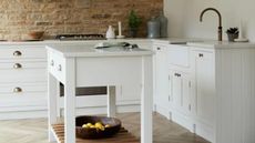 A clean ant-free white kitchen with a small kitchen island with an open wooden shelf, white kitchen cabinets around it, and a brown brick wall