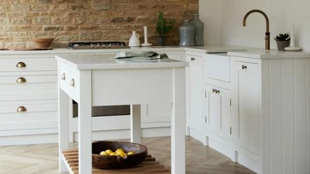 Portable kitchen island ideas like this small one with open sides and wooden shelving are so useful. Behind, a brick feature wall can be seen along with white cottage style cabinets can be seen with bronze fixtures. 