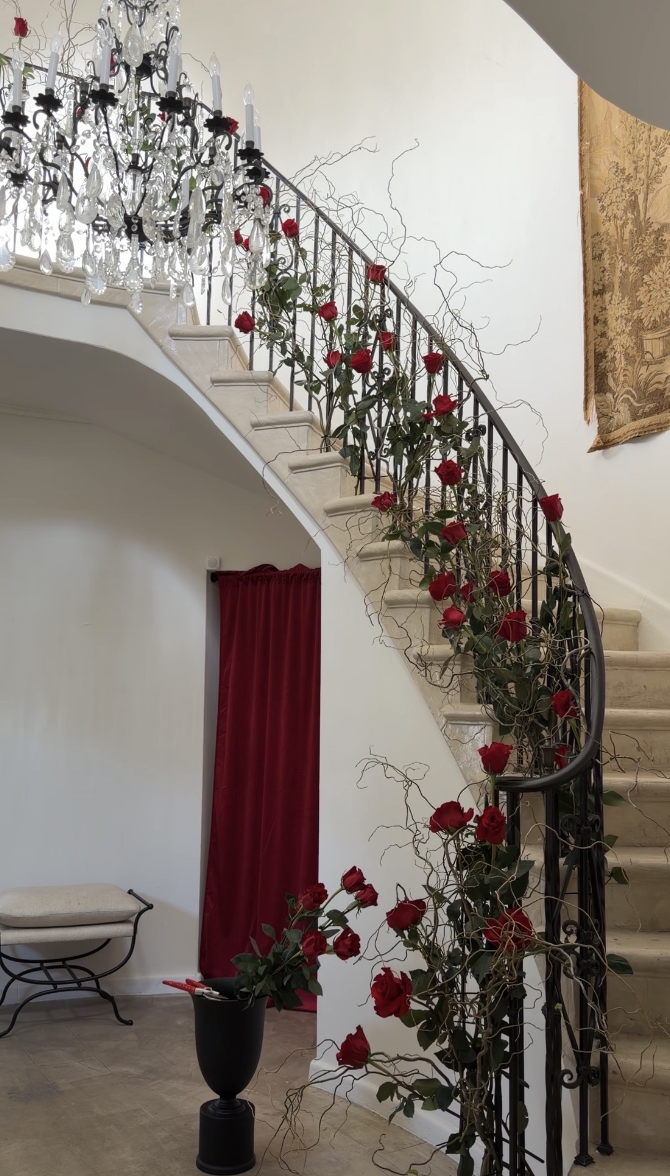 A spiral staircase adorned with red roses and branches with a red velvet curtained doorway underneath the staircase, a hallway bench, a chandelier and a large tapestry on the white walls