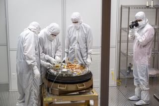 four people in clean room suits standing around a spacecraft capsule with an open top. one person, at far right, takes a picture