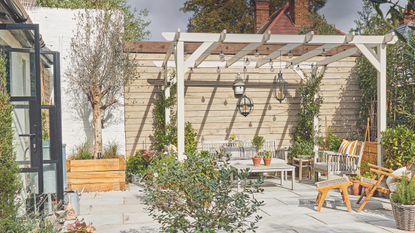 Patio area with wooden table, chairs and bench and plants under white wooden pergola in garden