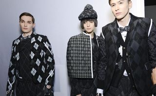 Three models wearing looks from Moncler Gamme Bleu's collection. They are wearing shirts, bow ties, jackets, gloves and a short, structure cape style piece in black, grey and white with a repeating diamond pattern
