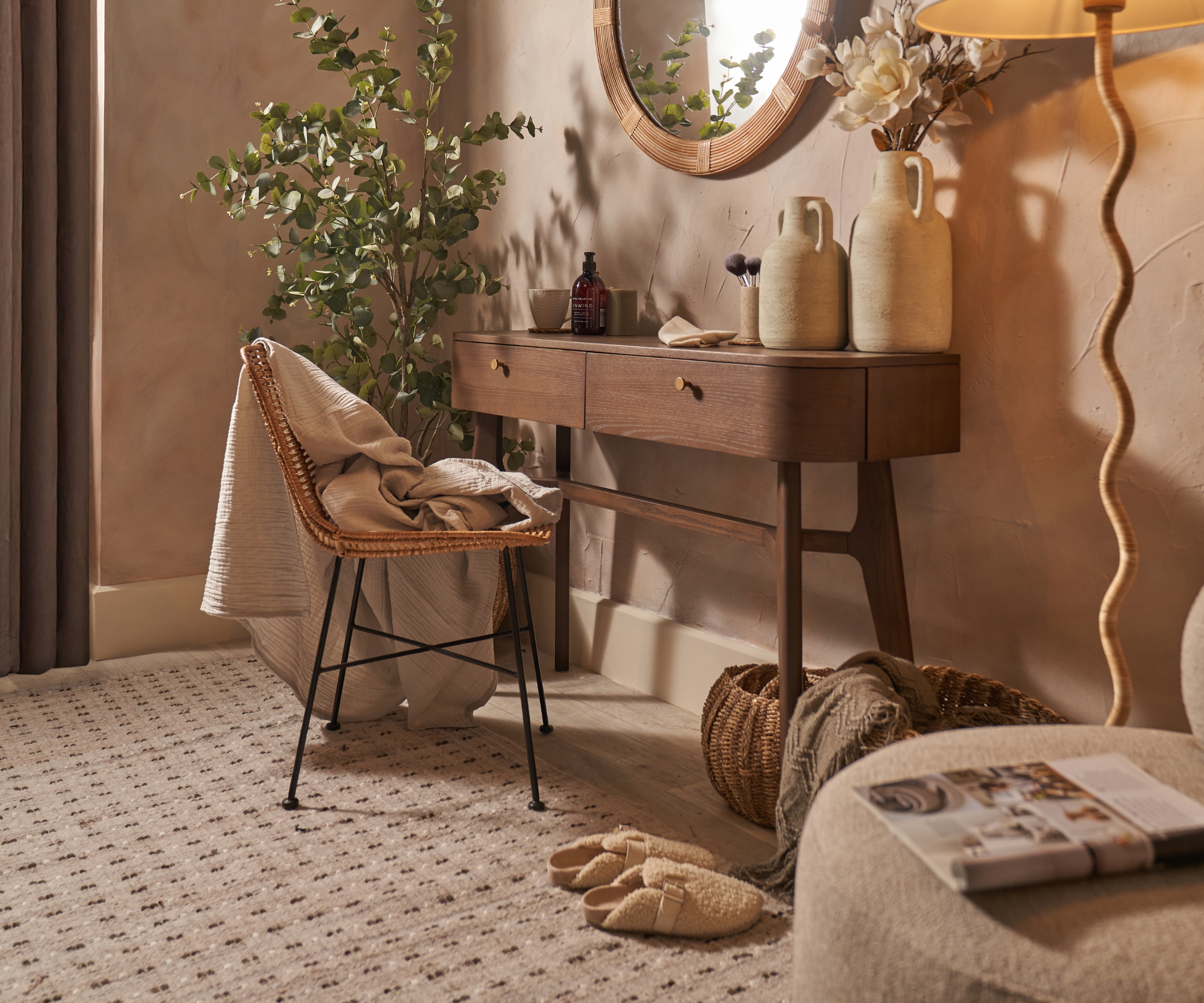 Wicker chair with throw beside a wood console table, oval mirror, vases, eucalyptus plant, and warm lamp light.