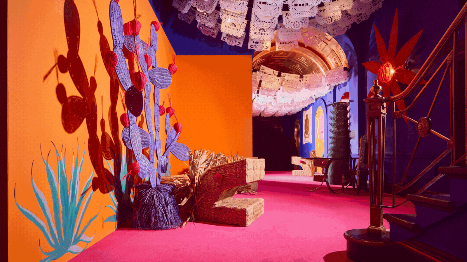 A rotating gif showcasing an indoor art installation characterized by bright hues of purple, orange, pink, and red across ginormous cacti-shaped wicker sculptures, animalesque creatures, and colorful ceilings.