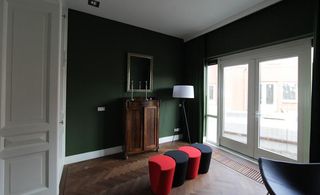 A room with dark green walls, dark wooden floor and white french doors. In the corner of the room is a white floor lamp and in the middle are 4 stools(2 red and 2 black)