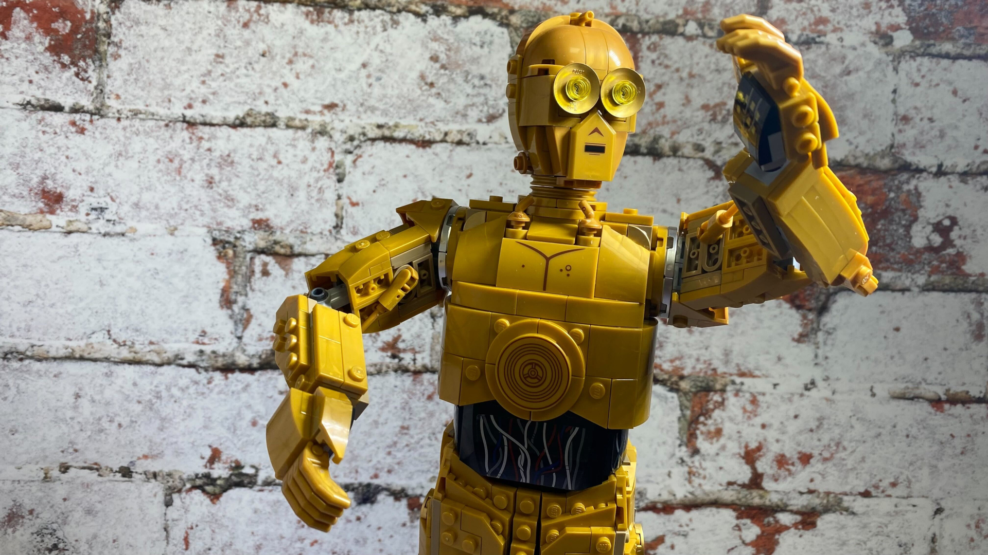 Lego C-3PO holding up his arm against a brick wall