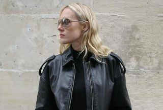 Woman wearing black turtleneck, black leather jacket, and sunglasses standing against Parisian wall.