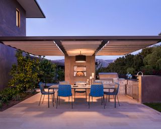An outdoor kitchen and dining area under a modern concrete pergola, lit with ambient lighting at twilight.