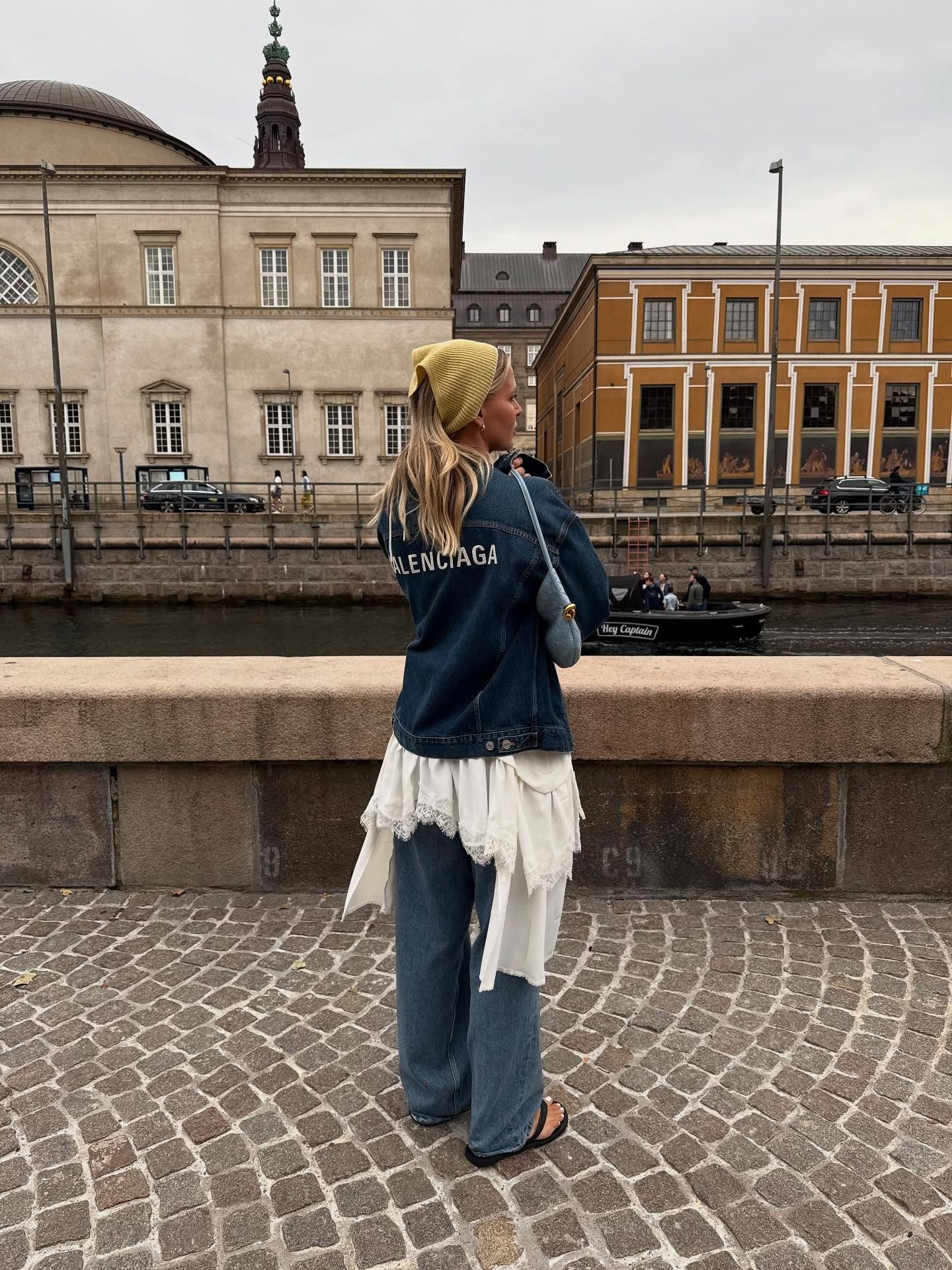 Fashion person Hollie Mercedes wears a Boho outfit. She wears a denim jacket, a layered lace dress, denim jeans and flip flops. She finishes the look with a lime green headscarf.