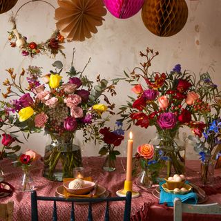 a table with pink tablecloth and an assortment of flower arrangements with pink and colourful tones candles on the table and hanging paper decorations