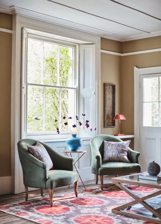 ochre living room with two emerald green armchairs, red and lilac rug, red table lamp, glass and wood coffee table, blue vase with cosmos on side table, traditional shutters on windows