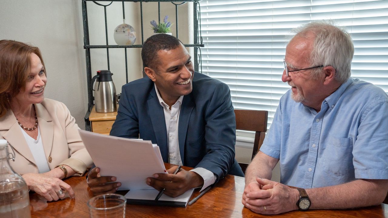 A financial planner smiles as he speaks with an older couple at their kitchen table.