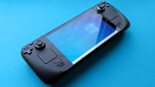 Steam Deck handheld gaming console with a black design is displayed against a blue background. The screen shows a blue Windows logo wallpaper