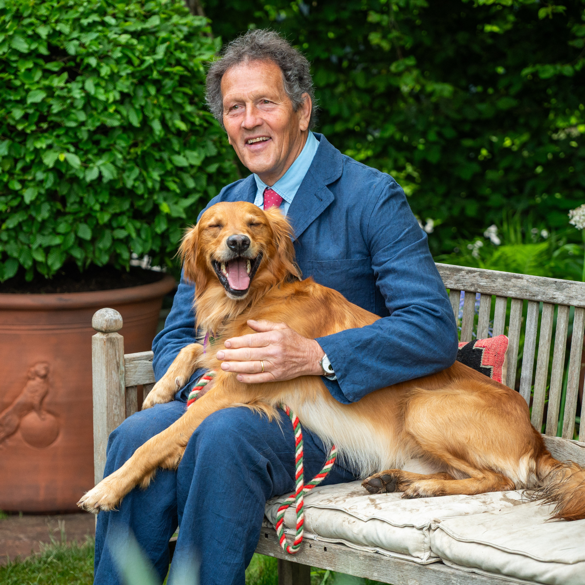 Monty Don sat on bench wearing blue suit. His golden retriver is sat on his lap.