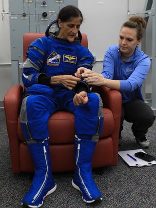 an astronaut sitting in a chair wearing a spacesuit. beside her a woman helps the astronaut with something on the arm. a panel is behind them