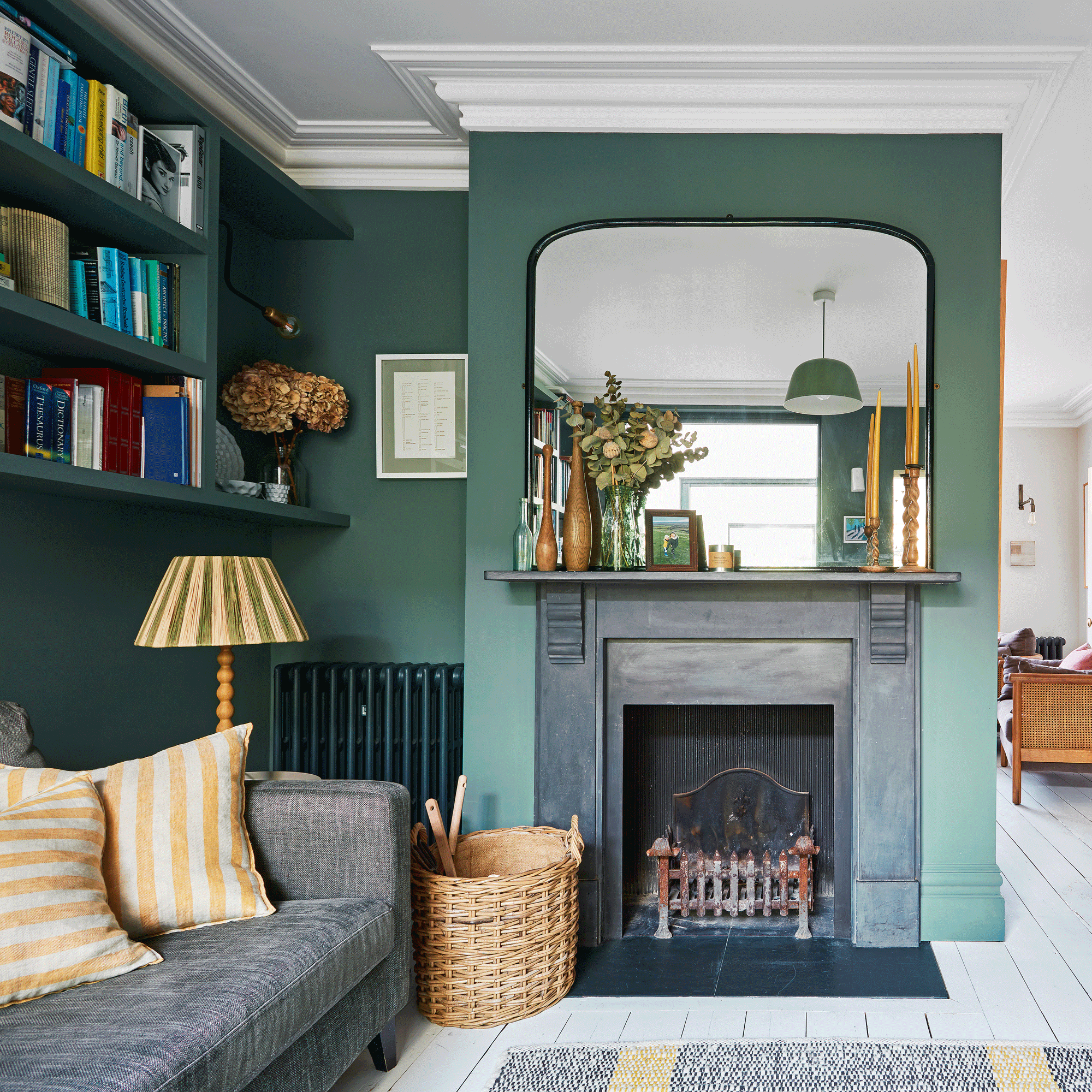 a living area with dark green walls and fitted shelving, a working fireplace with an overmantel mirror leading into another room