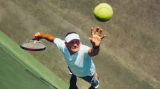 An older man or young retiree throws a tennis ball up to serve. The camera is above him and the ball.