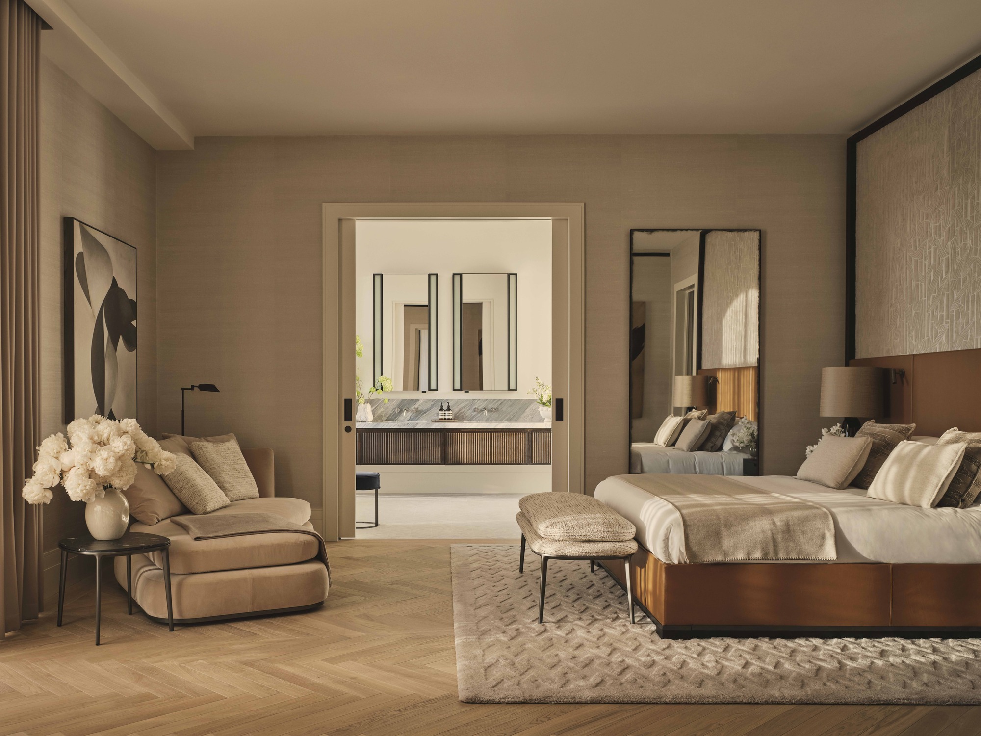 modern bedroom with herrigbone floorboards, upholstered bed with bench seat at the foot of the bed, daybed on the opposite wall with side table with vase of flowers, floor lamp and artwork, and doorway leading through to the bathroom