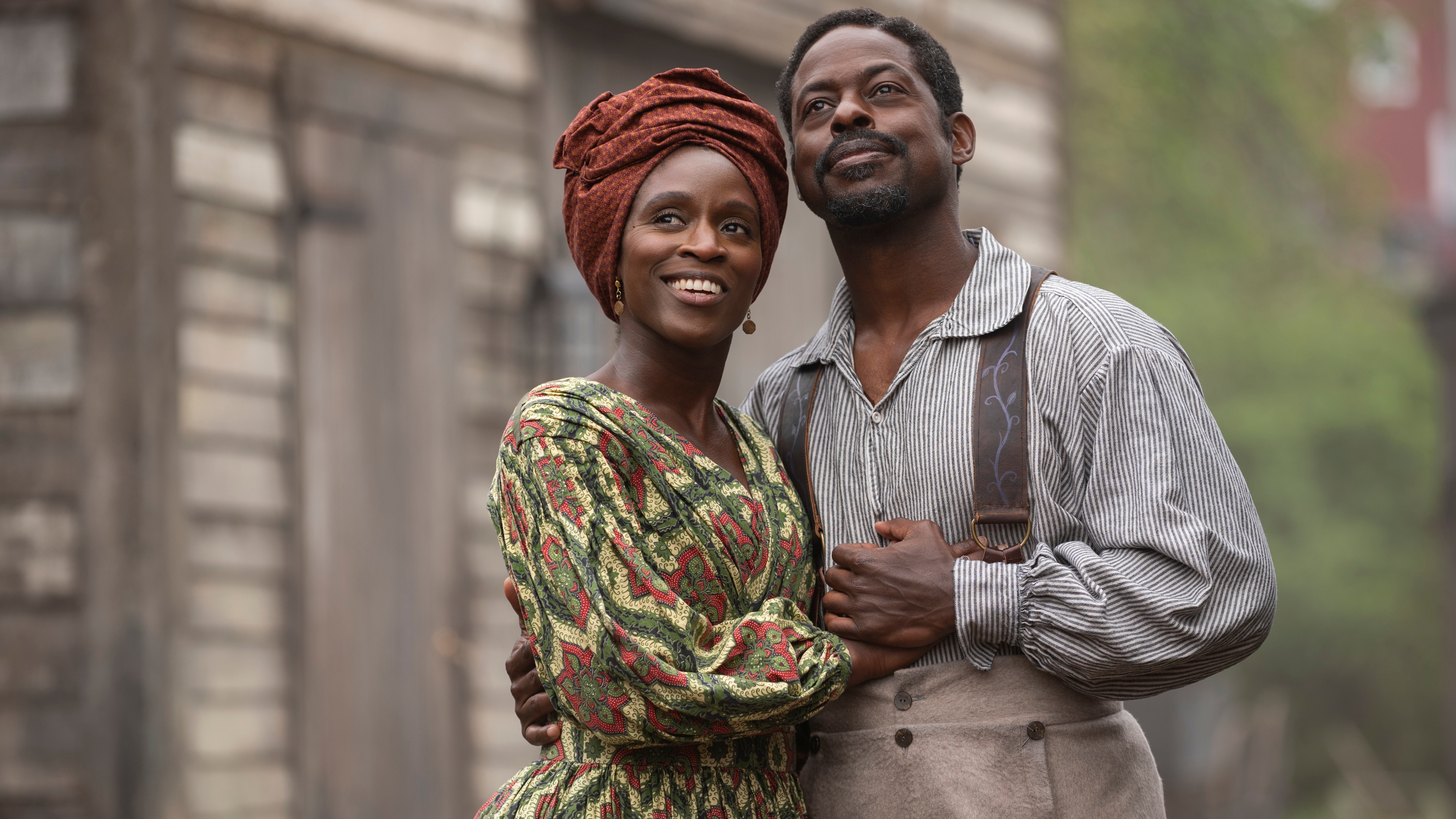 Sterling K Brown as Medwin and Sharon Duncan-Brewster as Miss Angie, a Trinidadian storeowner rebuilding her life in Nova Scotia and a maternal figure to Washington Black in times of trouble. Both actors are wearing 19th century period clothing, clasping each other and smiling at something in the distance.
