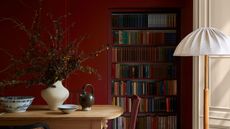 dining room with red painted walls, a built-in bookcase, floor lamp, wall with molding, timber dining table decorated with vase of flowers, teapot, bowl, and large bone china bowl