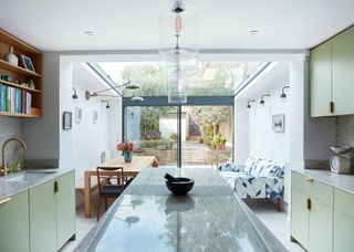 A glass ceiling kitchen extension with lights on either side of the wall. On one side of the kitchen is a large island with light pendants above it. There are also sage green upper and lower cabinets.