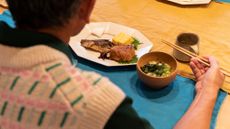 A view over someone's shoulder looking at their food on a table. They wear a white, pink and green patterned sweater and are holding chopsticks