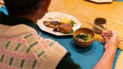 A view over someone's shoulder looking at their food on a table. They wear a white, pink and green patterned sweater and are holding chopsticks