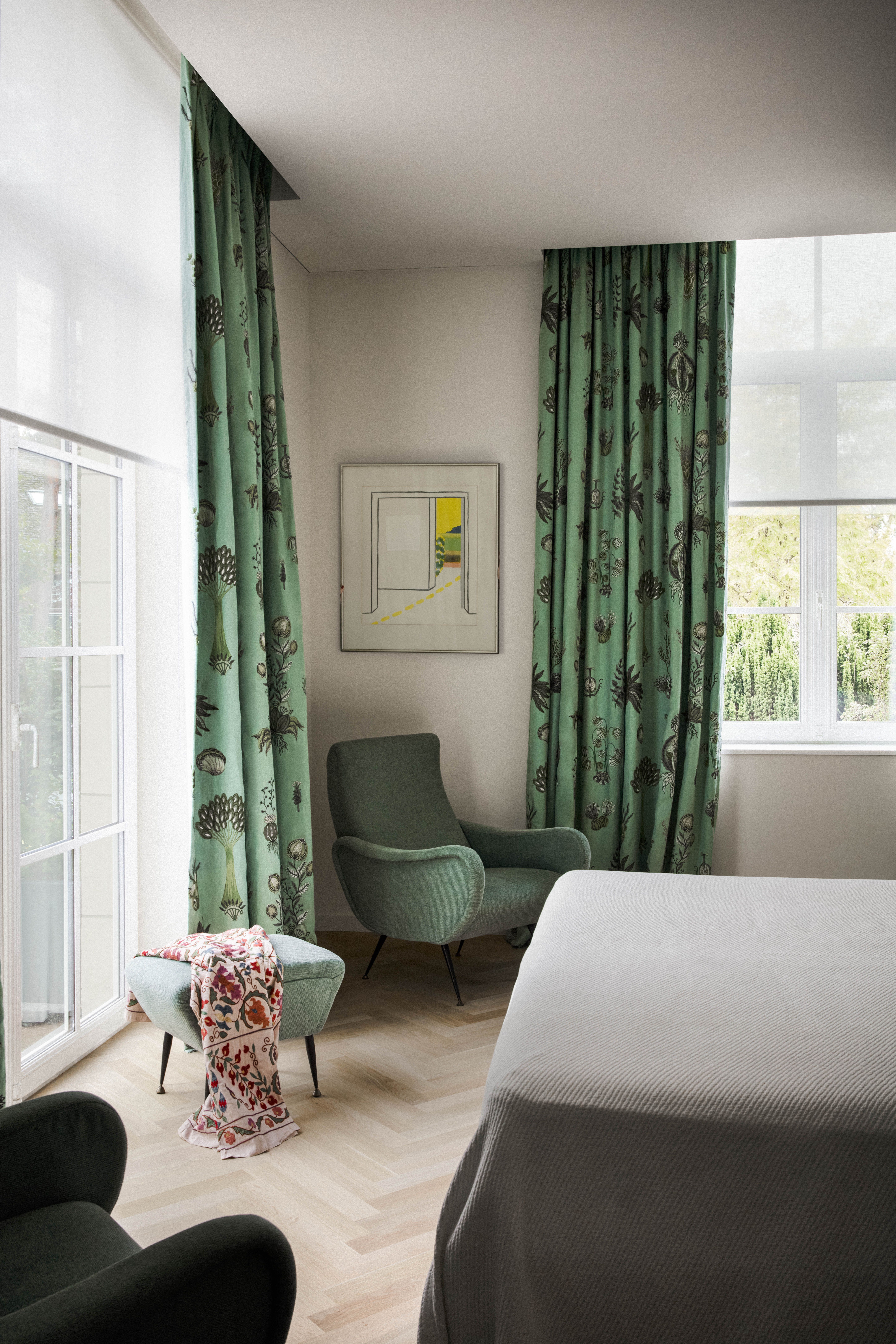 A bedroom in green tones, with a velvet headboard, printed green curtains and wooden flooring