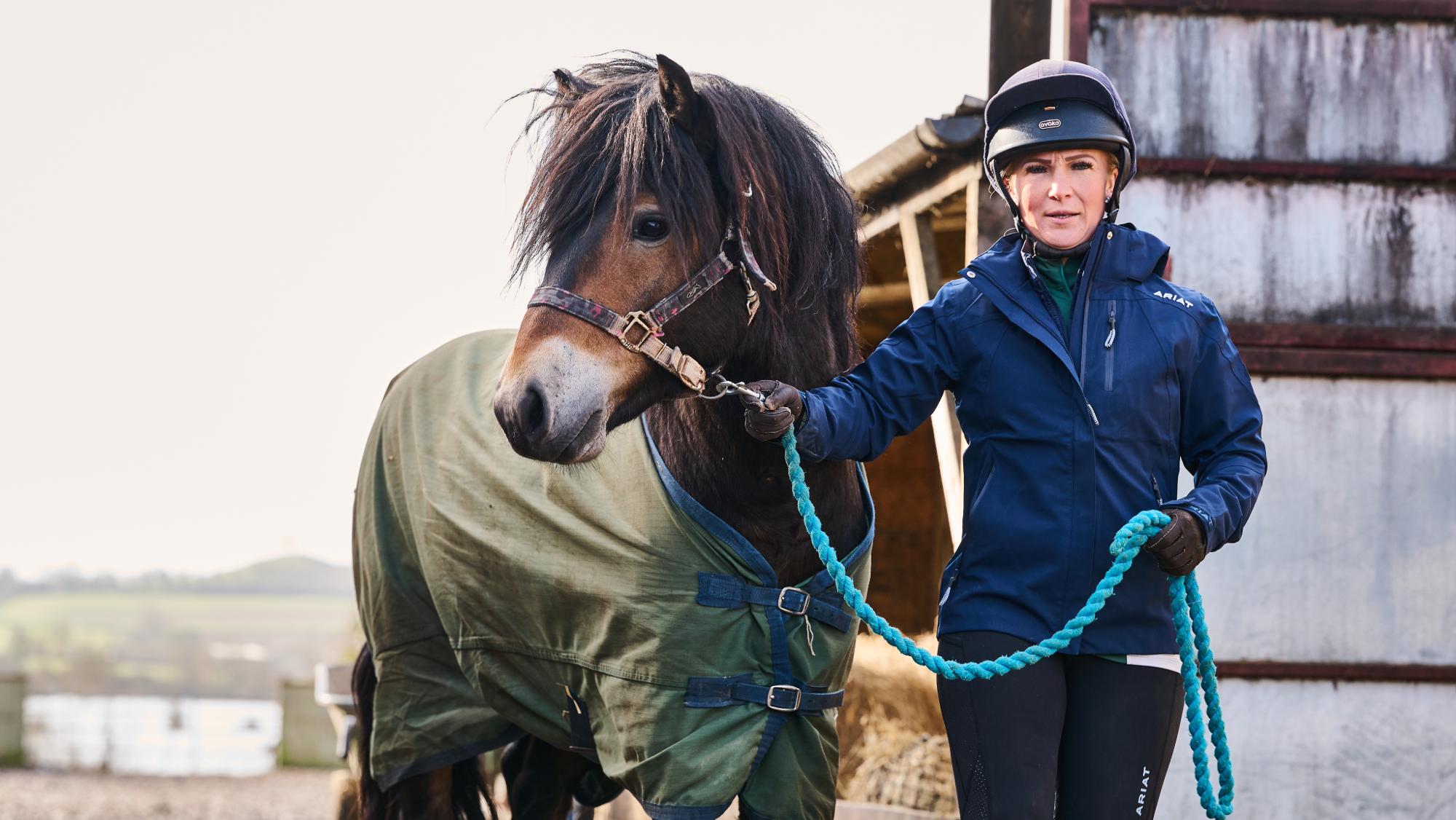 Woman in a navy blue jacket and wearing a hard hat leads a bay pony in a green rug
