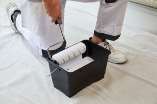 Person in white overalls kneeling down on clean white dust sheets with roller in hand, adding white paint from a black paint scuttle