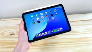 A hand holding up the iPad Mini 7 with Apple Pencil Pro attached in front of a wood table