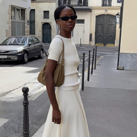 Woman wearing white cashmere t-shirt, white maxi skirt, brown bag, and black oval sunglasses on the streets of Paris.