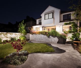 A view of the back of the house with wall lights and lighting in the flower beds with decorative paving and a shaped lawn