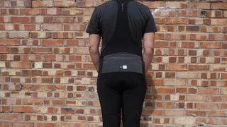 Rear view of rider standing in front of a wall wearing Pedaled Odyssey Women's Cargo Bib Tights